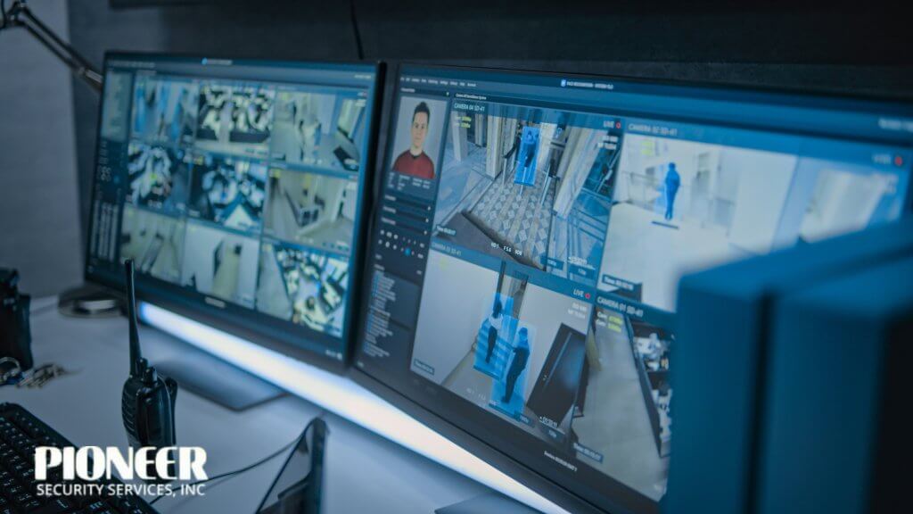 A close-up view of two computer monitors in a security control room. The screens display multiple live surveillance feeds from an office environment, featuring AI-powered person detection with bounding boxes, facial recognition on one corner, and security data. A two-way radio is visible on the desk.