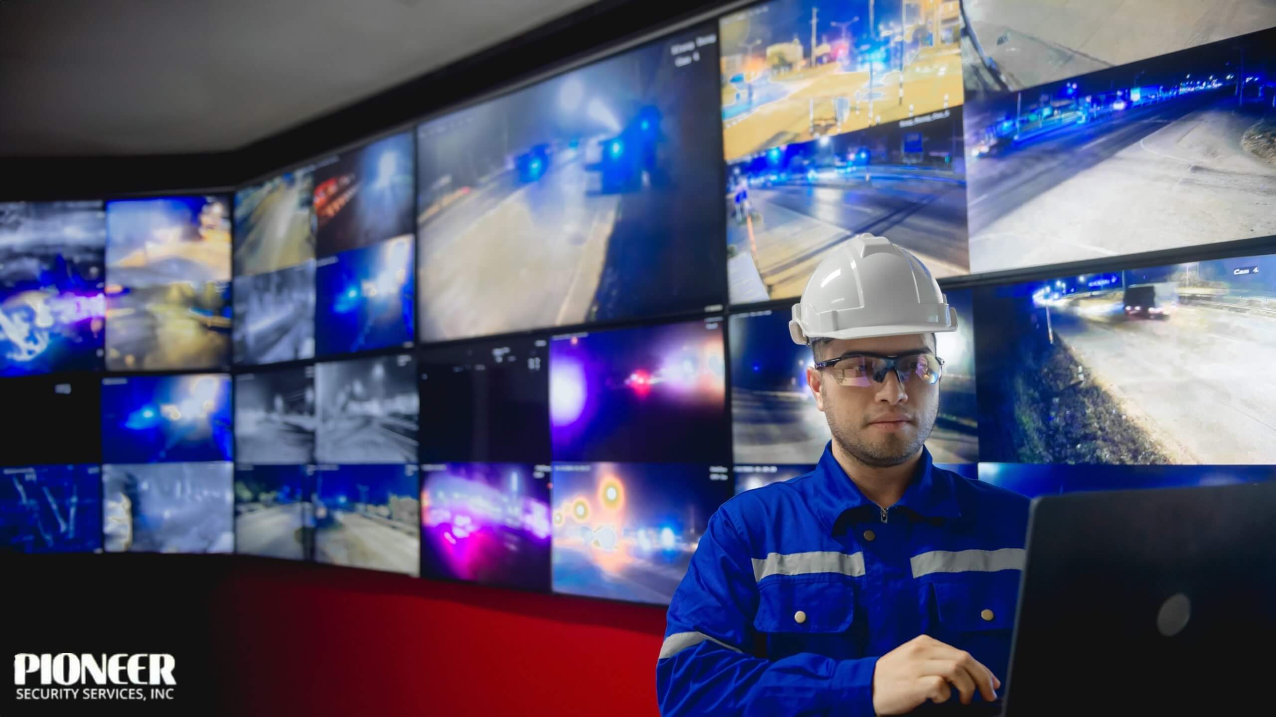 A man in a blue jumpsuit, white hard hat, and safety glasses works on a laptop in a control room. Behind him, a massive video wall displays a grid of low-light, blue-tinted surveillance footage of highways and city streets at night.