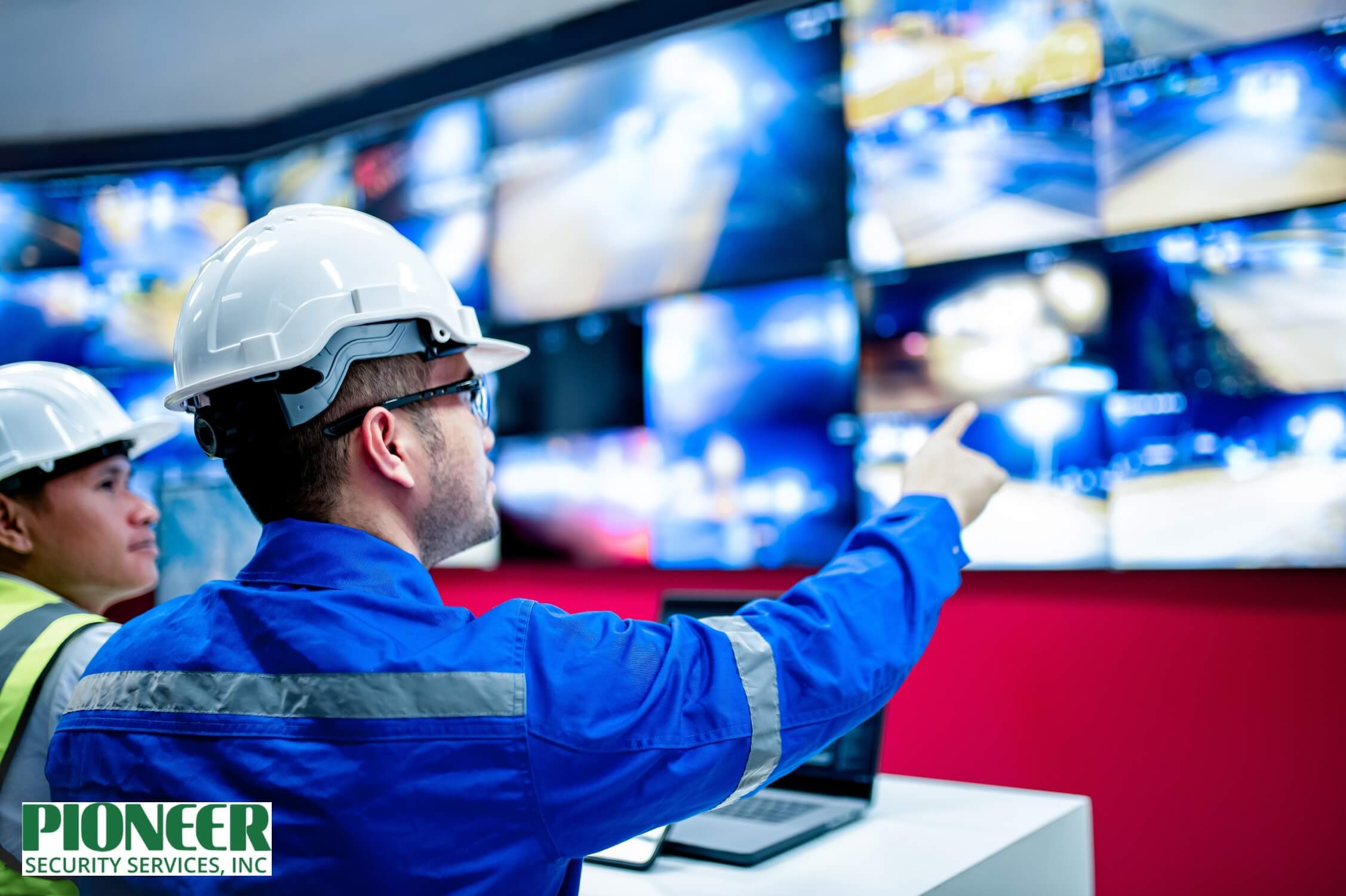 Two male engineers in hard hats and safety vests monitoring a large wall of blurred security camera screens, with one pointing to a view in a technical control room environment.