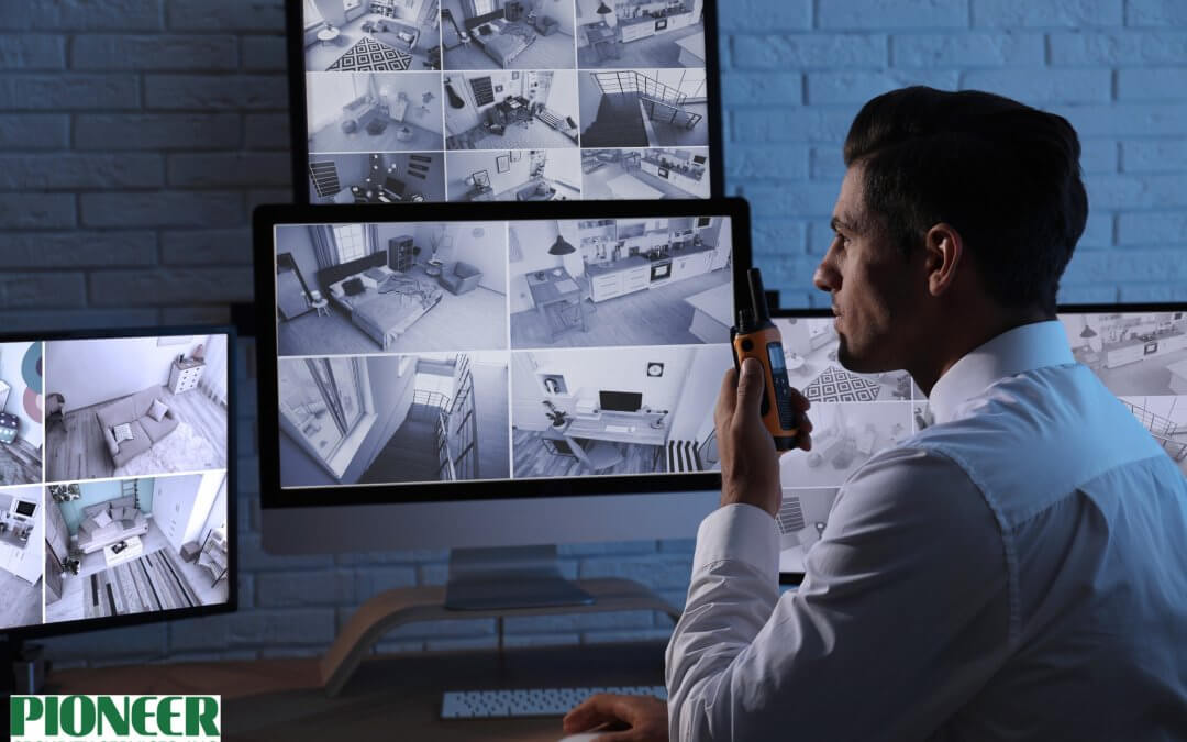 Security guard in a white shirt watching multiple live feeds from CCTV cameras on several dark-lit monitors, holding a two-way radio/walkie-talkie for communication.