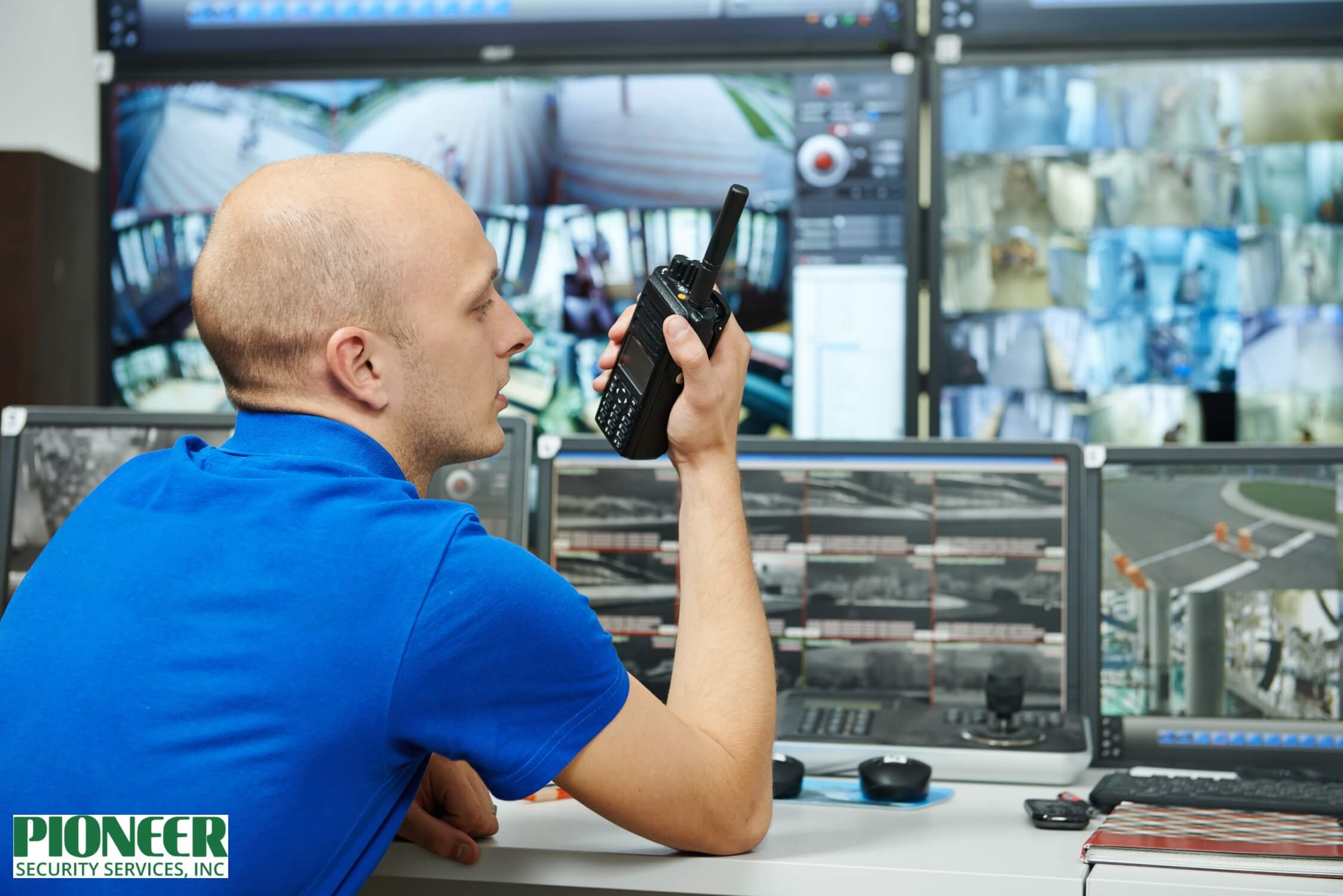 A male security operator wearing glasses and a headset sits in a dark industrial control room, focused on his computer. To his right, a large bank of monitors displays a grid of blue-tinted surveillance footage from an industrial or factory setting.