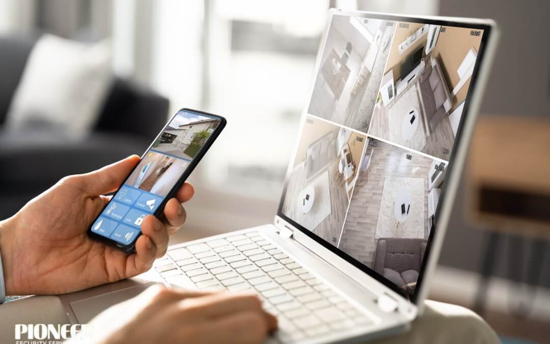 A person is monitoring home security on two devices in a bright living room. The laptop displays a four-panel grid of indoor surveillance feeds, while the person holds a smartphone showing a single outdoor camera feed along with smart home control buttons.
