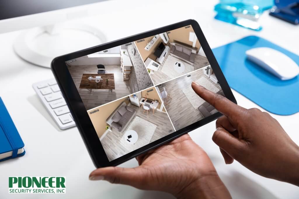 Close-up of hands holding a black tablet displaying four live security camera feeds from an interior office or home, emphasizing remote video surveillance access.