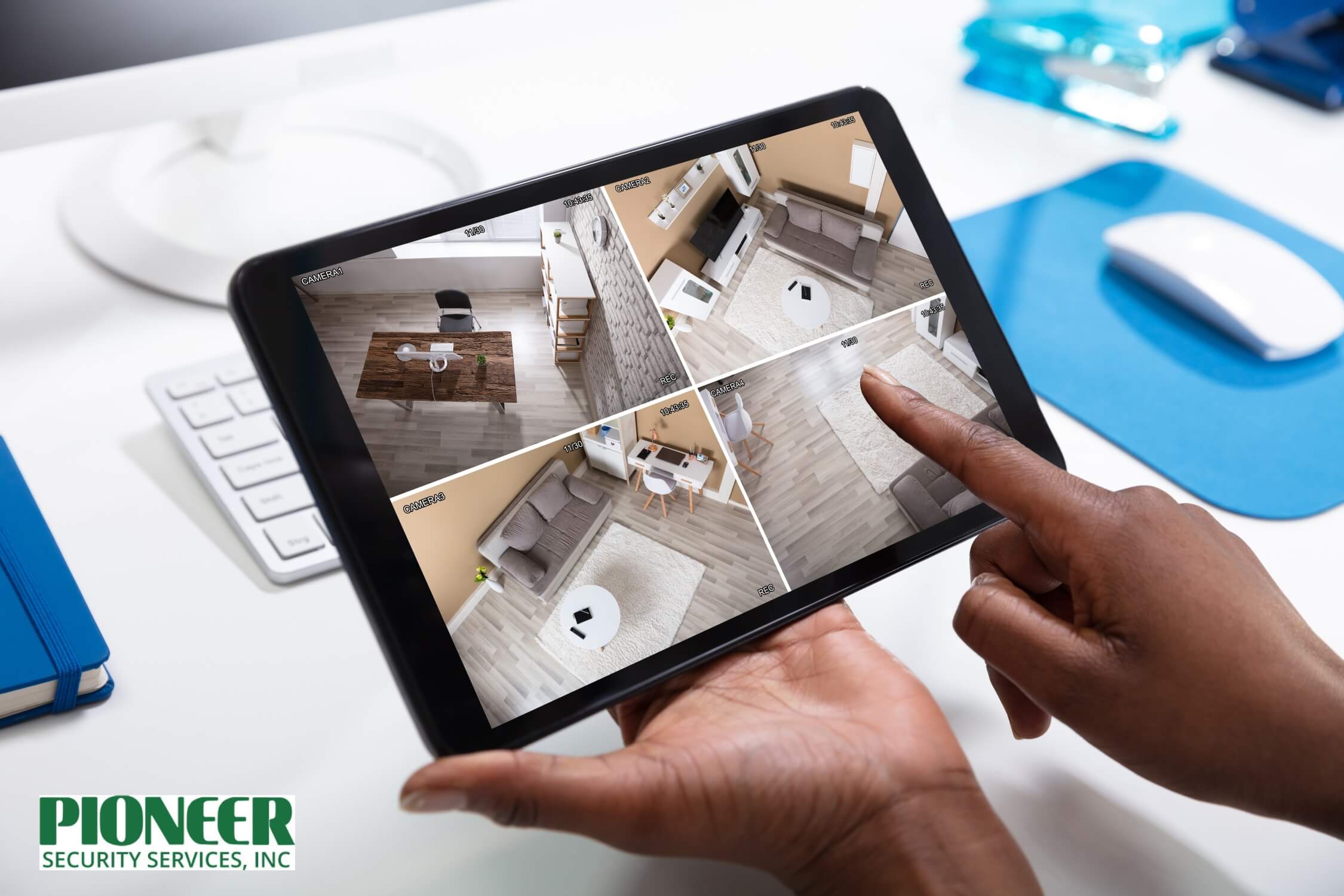 Close-up of hands holding a black tablet displaying four live security camera feeds from an interior office or home, emphasizing remote video surveillance access.