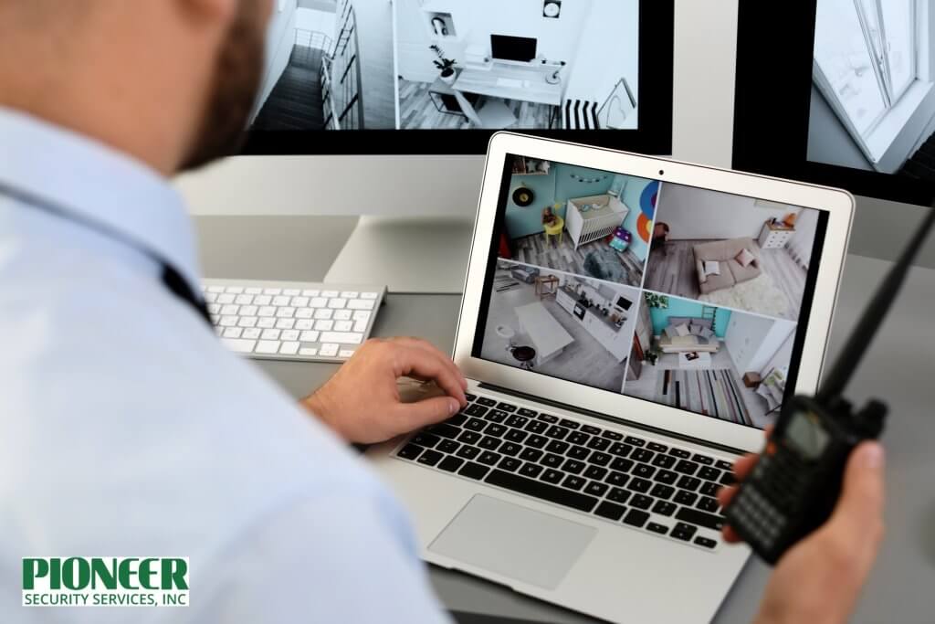 Over-the-shoulder view of a man in a security role looking at surveillance camera feeds on a laptop and a desktop monitor. The laptop screen shows a four-panel view of home interiors (kitchen, living room, nursery), and the man holds a two-way radio in his right hand.