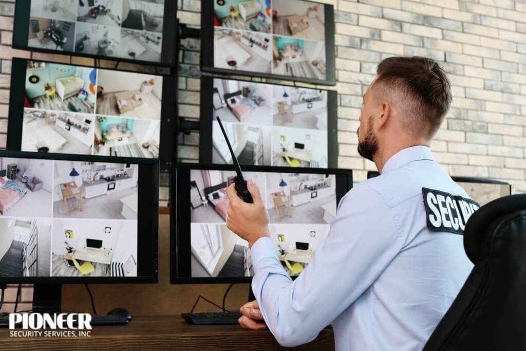 A security guard with a beard, wearing a light blue shirt with a "SECURITY" patch, sits with his back to the camera, holding a two-way radio. He is monitoring several screens displaying a grid of surveillance footage from various rooms inside a home or building.