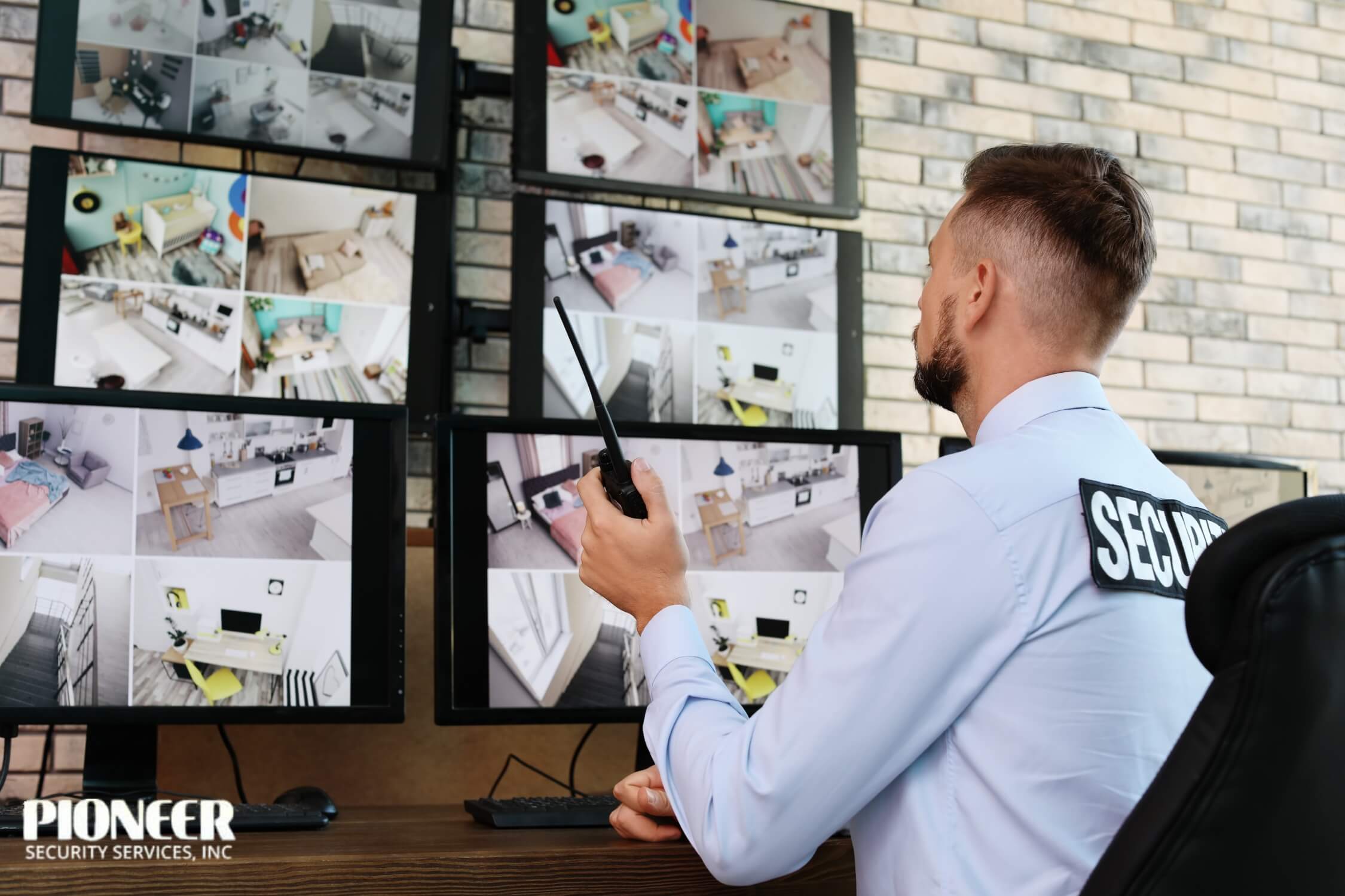 A security guard with a beard, wearing a light blue shirt with a "SECURITY" patch, sits with his back to the camera, holding a two-way radio. He is monitoring several screens displaying a grid of surveillance footage from various rooms inside a home or building.