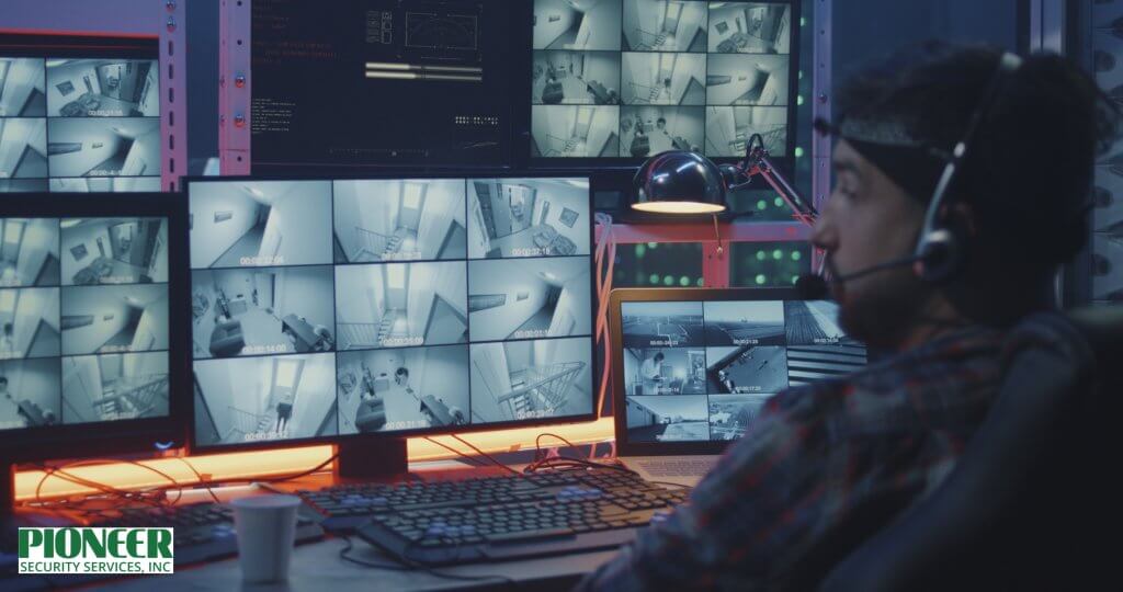 A security guard, viewed from behind, sits in a dark control room, wearing a cap and a white shirt. He is holding a two-way radio to his mouth and pointing at the grid of surveillance feeds displayed on his laptop, with a large wall of brightly lit security monitors behind him.