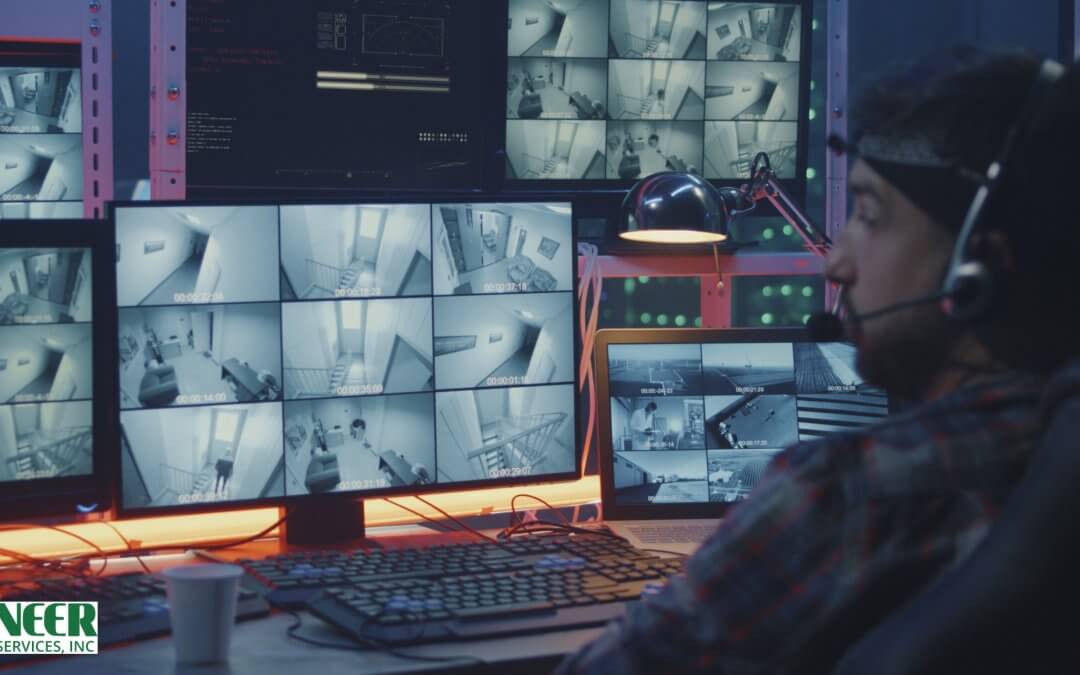 A security guard, viewed from behind, sits in a dark control room, wearing a cap and a white shirt. He is holding a two-way radio to his mouth and pointing at the grid of surveillance feeds displayed on his laptop, with a large wall of brightly lit security monitors behind him.
