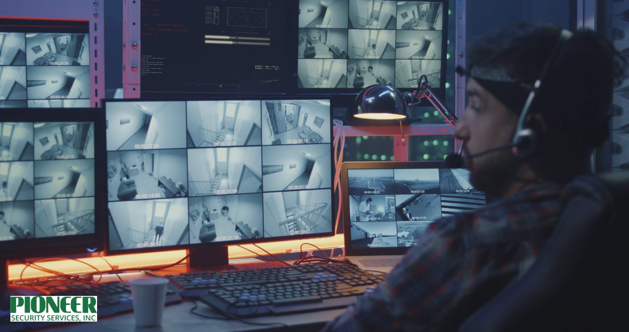 A security guard, viewed from behind, sits in a dark control room, wearing a cap and a white shirt. He is holding a two-way radio to his mouth and pointing at the grid of surveillance feeds displayed on his laptop, with a large wall of brightly lit security monitors behind him.