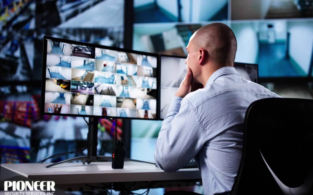 A bald man in a light blue shirt sits at a desk in a security control room, looking intently at a multi-monitor setup. One monitor displays a grid of many small surveillance video feeds, mostly showing hallways and stairwells, while a two-way radio sits on the desk in front of him.