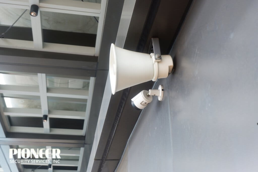 Close-up of a white horn loudspeaker mounted above a small white security CCTV camera on a dark gray wall of a contemporary building facade with large windows.
