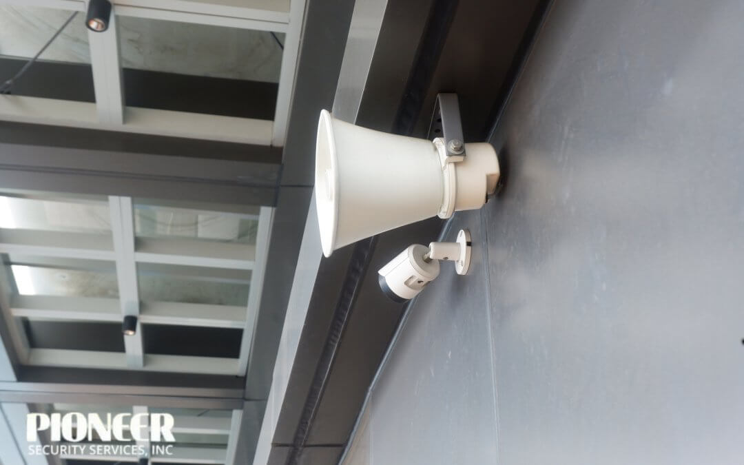 Close-up of a white horn loudspeaker mounted above a small white security CCTV camera on a dark gray wall of a contemporary building facade with large windows.