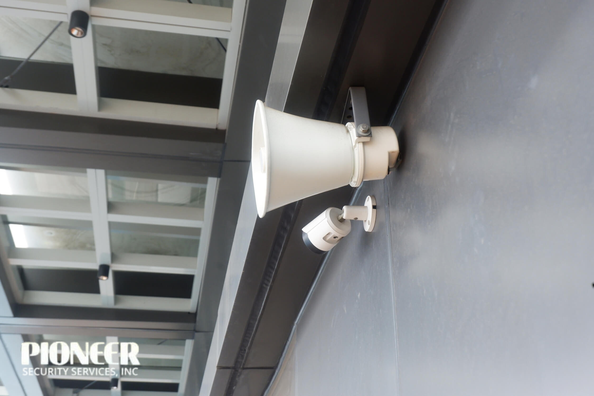 Close-up of a white horn loudspeaker mounted above a small white security CCTV camera on a dark gray wall of a contemporary building facade with large windows.