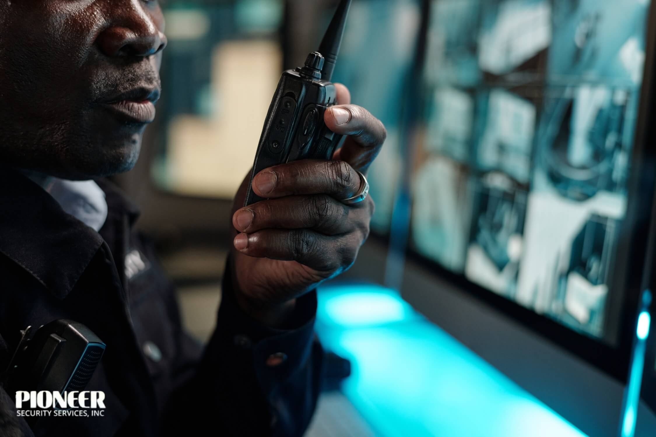A close-up shot of a Black security officer's face and hand as he speaks into a two-way radio. In the background, a computer monitor displays a grid of blue-tinted surveillance video feeds, indicating he is communicating while on duty in a control room.